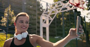 Güzel, mutlu, beyaz bir kızın açık havada akıllı telefondan gülümseyen ve selfie çeken portresini yakından çek. Neşeli, genç, güzel bayan sporcu basketbol sahasında cep telefonuyla fotoğraf çekiyor..