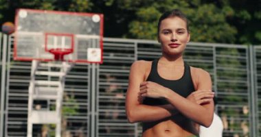 Güneşli bir günde dışarıda basketbol sahasında kameraya gülümseyen neşeli genç kadın portresi. Erkek arkadaşları sokak basketbolu çalışırken mutlu, güzel beyaz kız ayakta duruyor.