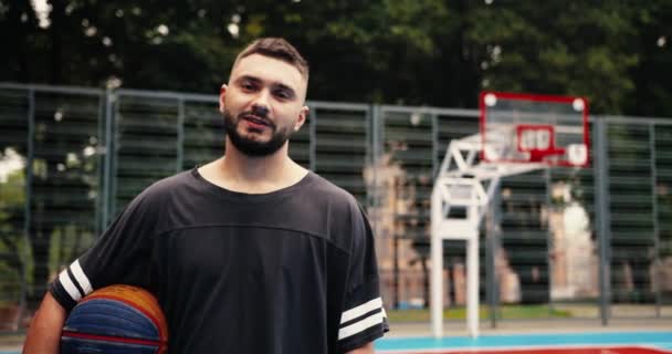 Gros plan portrait d'un heureux athlète masculin caucasien debout sur une aire de jeux urbaine et tenant le ballon à l'extérieur dans la rue. Beau joueur de basket regardant la caméra. Afro-américain jouer sur fond — Video