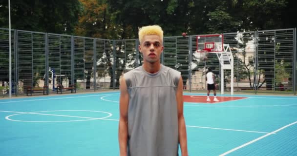 Retrato de deportista serio con cabello rubio parado en cancha de baloncesto al aire libre en la ciudad. Streetball jugador mirando a la cámara en el patio de recreo. Mixed-carreras chicos jugando en segundo plano. — Vídeo de stock