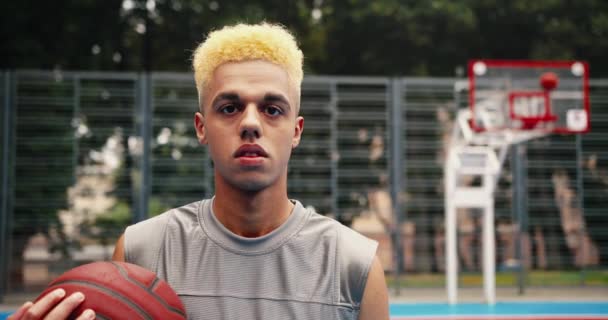 Gros plan portrait de jeune homme sérieux debout sur le terrain de basket urbain avec balle dans les mains en plein air. Beau joueur de streetball masculin au visage calme regardant la caméra sur la rue Concept de style de vie sportif — Video