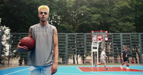 Joven niño alegre en ropa deportiva de pie en el patio de recreo urbano al aire libre con pelota. Alegre jugador de baloncesto masculino sonriendo a la cámara en la calle mientras sus amigos practican streetball. Concepto atlético — Vídeo de stock