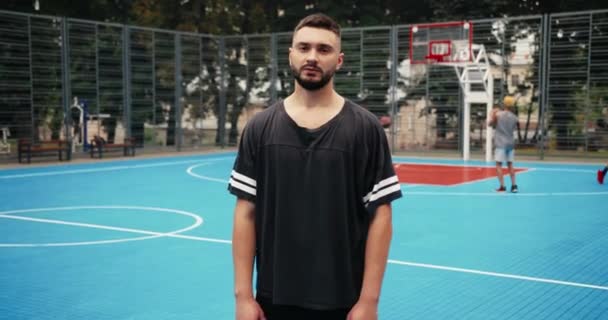 Junger ernsthafter kaukasischer Typ in Sportkleidung, der auf einem städtischen Spielplatz steht. Nahaufnahme Porträt eines männlichen Basketballspielers, der auf der Straße in die Kamera schaut, während seine Freunde Streetball praktizieren — Stockvideo