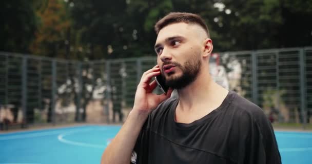 Primer plano retrato del joven hombre caucásico de pie en la cancha de baloncesto urbano al aire libre y charlando en el teléfono inteligente. Un jugador serio de streetball hablando por teléfono móvil en la calle. Concepto de llamada — Vídeo de stock