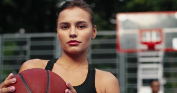 Közelkép komoly, fiatal, csinos, kaukázusi női atléta kosárlabdára néző kamerával, miközben kint áll a játszótéren. Gyönyörű, nyugodt arcú nő portréja. Atlétikai koncepció — Stock videók