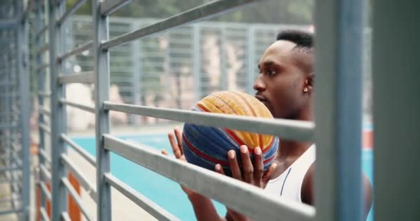 Портрет красивого молодого афроамериканского спортсмена, стоящего на открытой городской площадке для стритбола и бросающего мяч между руками и отводящего взгляд. Крупный план серьезного баскетболиста в городе — стоковое видео