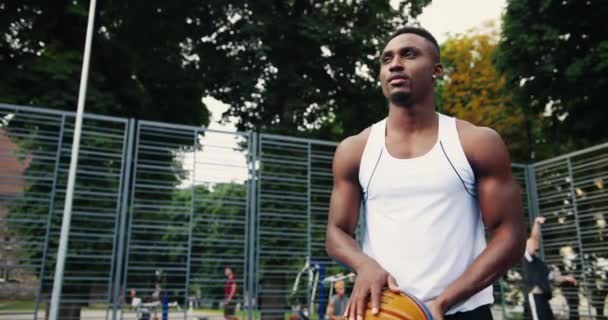 Portrait d'un beau jeune joueur de basket-ball professionnel afro-américain pratiquant sur une aire de jeux extérieure et lançant une balle. Homme caucasien jouant sur fond. Concept de formation — Video