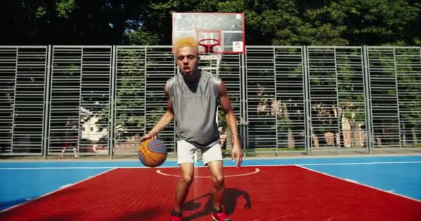 Boldog fiatal férfi kosárlabda játékos sportruházat csöpögő labda, gyakorló egyéni készségek kezelése és pontozási labda a karika álló háttal kosár a szabadtéri kosárlabda pályán — Stock videók