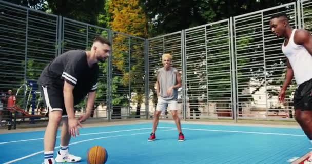 Aktiva atletiska multietniska manliga vänner tränar streetball spel på utomhus basketplan. Kaukasiska och afroamerikanska män basketspelare tränar färdigheter. Bakre delen av killen gör mål. — Stockvideo