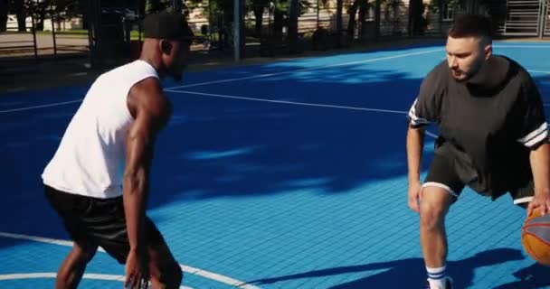 Multi-ethnic sportsmen playing basketball outdoors on sunny day. Caucasian male player training cross bouncing between legs and dribbling on local court. African American guy covering opponent — Stock Video