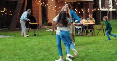 Beyaz mutlu anne ve baba bahçedeki yeşil çimlerde koşup çocuklarla oynuyorlar. Arka planda aile barbeküsü. Anne ve babayla neşeli çocuklar eğleniyor ve gülüyorlar. Dışarıda..