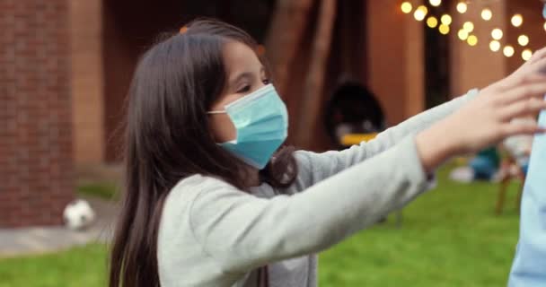 Close up of Caucasian small cute granddaughter in medical mask hugging her grandfather in yard. Barbeque meal on background. Little girl embracing grandpa in face shield outdoors. Pandemic concept. — Stock Video