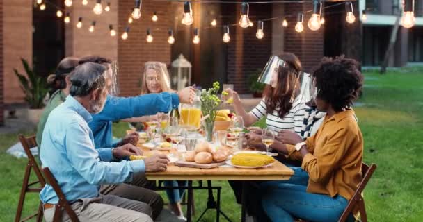 Mixed-races people in masks talking at dinner table in back yard. Multi ethnic family having picnic meal and chatting friendly. Communication of generations during pandemic. Pouring drinks and juice. — Stock Video