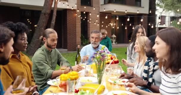 Multi etnikai család ül az asztalnál étkezés szabadtéri piknik és idősebb férfi mond tósztot. Vegyes versenyek boldog fiatal és idős emberek vacsoráznak és pirítós party barbrque Ünneplés hétvégén — Stock videók