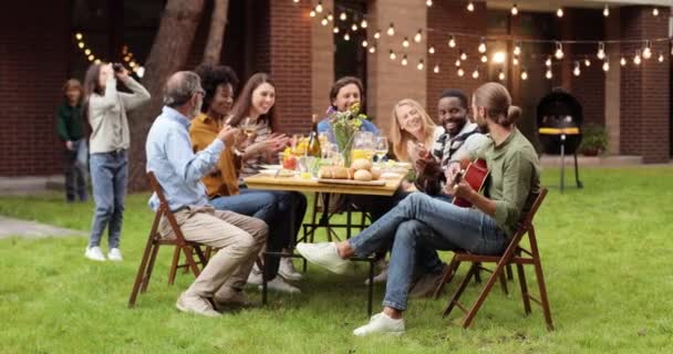 Mixtes-courses heureux grande famille assis à la table avec dîner à l'arrière-cour à l'extérieur et avoir du plaisir. Un type qui joue de la guitare devant des gens souriants et multiethniques. Musique. Musicien divertissant au pique-nique — Video