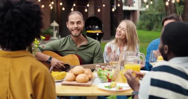 Erkek müzisyen akşam yemeği masasında ailesiyle gitar çalıyor ve şarkı söylüyor. Melez arkadaşlarla hafta sonu. Adam müzik çalıyor. Arka bahçede piknikte çok ırklı insanlar eğleniyor. barbekü — Stok video