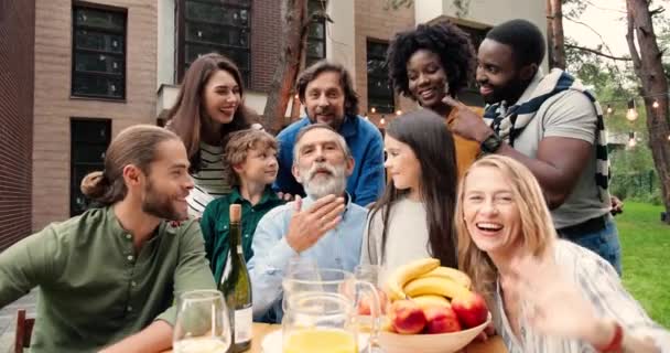 Glada multietniska familjemedlemmar ler och vinkar till kameran samtidigt som de äter lunch på bakgården vid bordet och videochattar. Videochatt för blandraser. Olika åldrar. Helg på picknick — Stockvideo