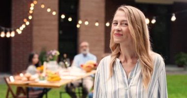 Beyaz, orta yaşlı, güzel sarışın bir kadının portresi avluda dikiliyor, yüzünü kameraya çeviriyor ve neşeyle gülümsüyor. Aile, arka plandaki yemek masasında. Mutlu güzel kadın