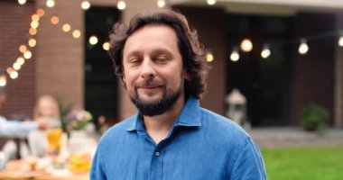 Bahçede durup kameraya gülümseyen beyaz sakallı adamın portresi. Aile barbeküsünde bir erkek. Arka planda yemek masasında oturan mutlu insanlar. Açık havada. Dolly vurdu. Yüzü yakınlaştırıyorum..