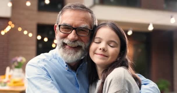Portrait of Caucasian grandfather in glasses hugging small cute granddaughter and smiling in yard. Old gray-haired man embracing little pretty girl. Celebration outdoors. Family dinner and meeting. — Stock Video