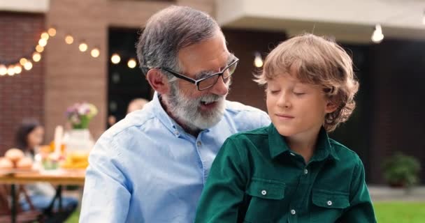 Portrait of Caucasian grandfather in glasses hugging small cute grandson and smiling in yard. Old gray-haired man embracing little boy. Celebration outdoors. Family dinner and meeting. Happiness. — Stock Video