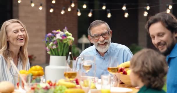 Keluarga bahagia Kaukasia yang ceria duduk di meja makan di luar pada akhir pekan dan menertawakan anak kecil. Orangtua dan kakek-nenek tersenyum dan makan bersama dengan anak laki-laki dan cucu perempuan. — Stok Video