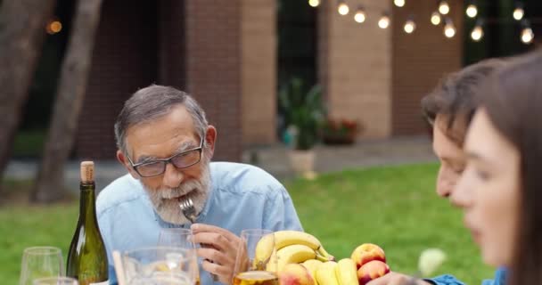 Az öreg kaukázusi nagyapa és apa együtt eszik és beszél gyerekekkel vagy unokákkal a ház udvarán hétvégenként. Idősek és fiatalok együtt vacsoráznak vagy ebédelnek a barbecue-n. — Stock videók