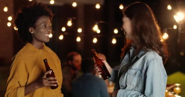 Dos mestizas jóvenes guapas y felices chicas mejores amigas hablando y riendo con botellas de cerveza en las manos en la fiesta por la noche. Tarde en la noche. Hermosa mujer charlando y sonriendo mientras bebe. — Vídeos de Stock