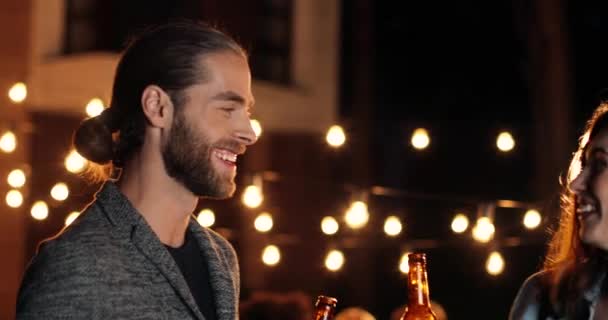 Caucásico joven feliz chica y chico, mejores amigos hablando y riendo con botellas de cerveza en las manos en la fiesta por la noche. Tarde en la noche. Hermosa mujer y hombre guapo charlando y sonriendo. — Vídeos de Stock