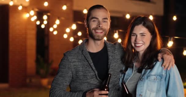 Retrato de un joven guapo y hermoso hombre y mujer caucásicos sonriendo alegremente y de pie en la fiesta exterior por la noche. Feliz chico y chica en la celebración de la noche. Pareja en abrazos. — Vídeos de Stock