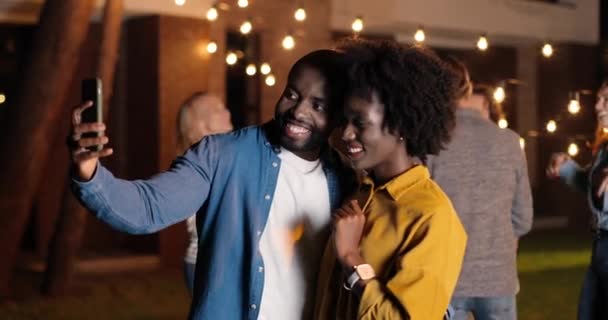 Afroamericano joven feliz y alegre pareja, hombres y mujeres sonriendo y posando en el teléfono inteligente mientras toma fotos selfie por la noche. Fiesta tardía. Hombre y mujer abrazándose y divirtiéndose. — Vídeos de Stock