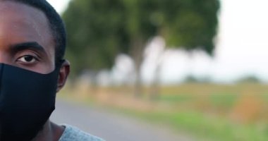 Spor yaptıktan ya da koştuktan sonra kameraya bakan maskeli yorgun, Afro-Amerikalı, terli bir sporcunun yüzünün yarısını kapatın. Açık havada terli erkek koşucunun portresi. Salgın. Covid 19.