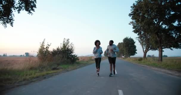 Afro-Amerikaanse jonge sportieve man en vrouw joggers rennen op de weg vroeg in de ochtend op het platteland. Een paar lopers. Sportvrouw en sportman joggen in de natuur. — Stockvideo