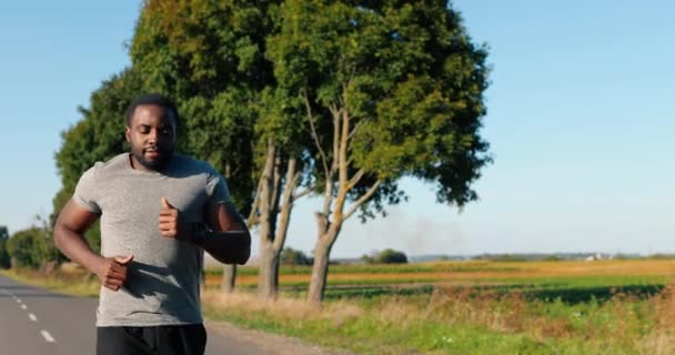 Afro-Amerikaanse knappe sterke mannelijke jogger rent op de weg op het platteland op de zomerdag. Aantrekkelijke sportieve man aan het joggen buiten. Sport concept. Sportman hardloper. Ochtendnatuur. — Stockvideo