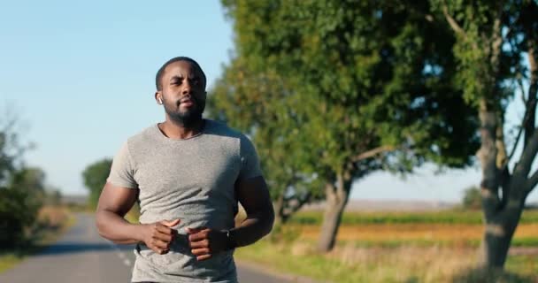 Afro-Amerikaanse knappe sterke mannelijke jogger in koptelefoon loopt op de weg op het platteland op de zomerdag. Aantrekkelijke sportieve man joggen buiten. Sport concept. Sportman hardloper. Ochtendnatuur. — Stockvideo