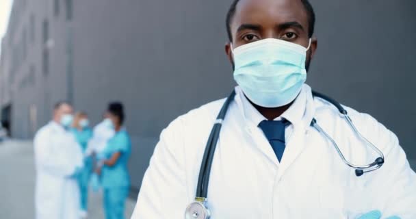 Retrato de un hombre afroamericano guapo doctor en máscara médica y con estetoscopio mirando a la cámara. Primer plano del médico masculino en protección respiratoria. Médicos de razas mixtas en segundo plano. — Vídeo de stock