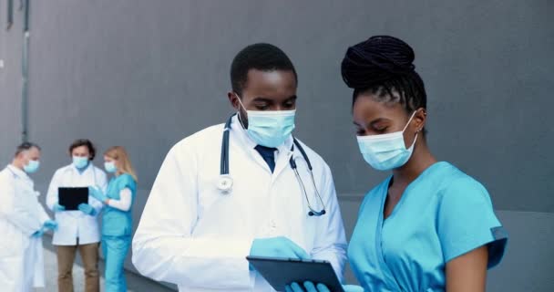 Pareja afroamericana de hombre y mujer, doctores colegas en máscaras médicas trabajando y usando un dispositivo de tableta. Médicos hombres y mujeres hablando, tocando y desplazándose en la computadora gadget. — Vídeos de Stock