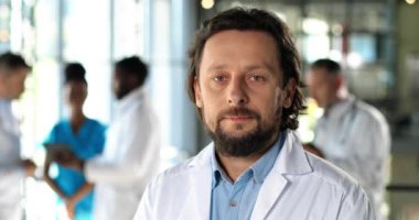 Kameraya bakıp klinikte dikilen beyaz, yakışıklı bir doktor. İçeride. Hastanedeki erkek doktor. Arka planda konuşan melez doktorlar. Beyaz önlüklü doktor portresi.