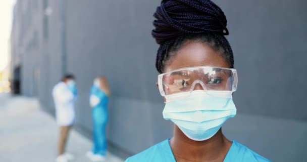 Retrato de afro-americano bela mulher médica em máscara médica e óculos olhando para a câmera. Close up médico feminino em proteção respiratória. Médicos em segundo plano. A aproximar-se. Dolly atirou — Vídeo de Stock