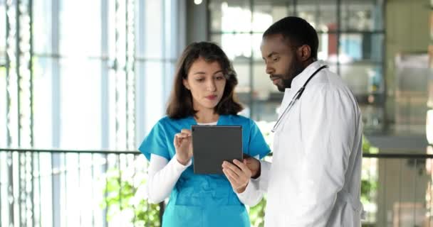 Mezclas de razas jóvenes pareja de hombres y mujeres médicos de pie en el hospital, hablando y utilizando el dispositivo de tableta. Multi étnico hombre y mujer, médicos discutiendo el trabajo y viendo en gadget. — Vídeos de Stock