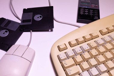 Eski bir bilgisayar klavyesinin üst açısını, kablolu bir fareyi, bazı disketleri ve beyaz bir arkaplanda uzaktan kumandayı göster.