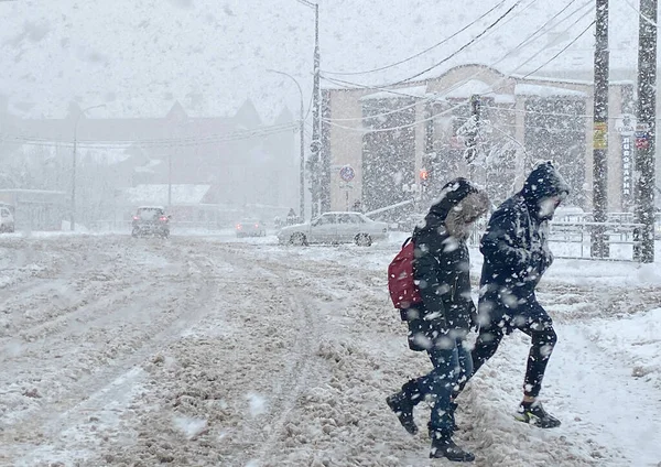 Снежная буря в городе. Молодая пара во время метели идет через улицу. Автомобили на снежной дороге. — стоковое фото