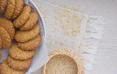 Beyaz tabakta susamlı kurabiyeler hasır sepette susam tohumları, pürüzlü masa örtüsü.