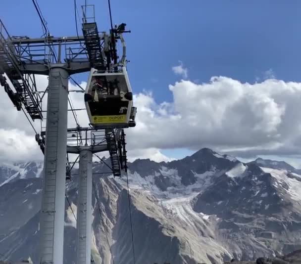 Gondola linbana på Elbrus. Gula stugor som rör sig upp och ner med hjälp av en linbana mot bakgrund av bergstoppar och en blå himmel med moln. — Stockvideo