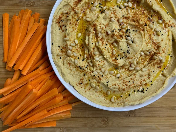 Hummus fait maison saupoudré d'huile d'olive, décoré de pignons de pin, avec des bâtonnets de carotte sur une assiette blanche. — Photo