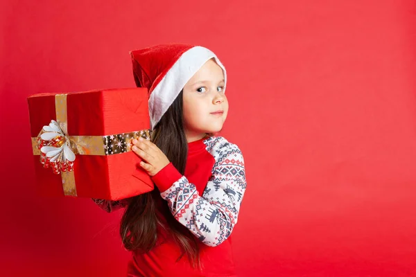 close up girls in pyjamas and a Santa Claus hat hold a gift box with a gold bow at the ear on a red background