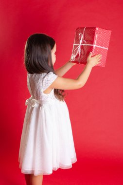 Beyaz elbiseli bir kız kırmızı bir hediye kutusu tutuyor, canlı bir arka planda izole edilmiş.