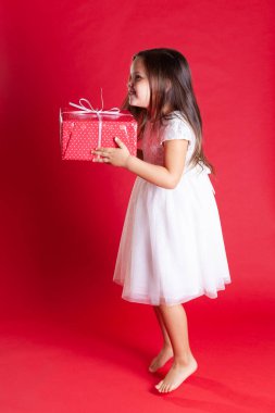 Beyaz elbiseli uzun boylu bir çocuk mutluluktan yana zıplıyor ve kırmızı arka planda izole edilmiş bir hediye kutusu tutuyor.