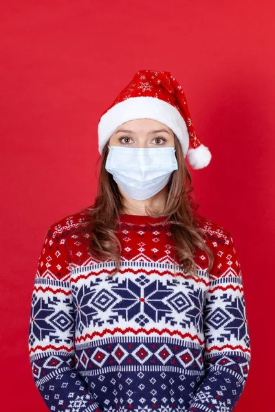 Mujer joven en máscara médica, sombrero de Santa Claus y suéter estampado aislado sobre fondo rojo —  Fotos de Stock