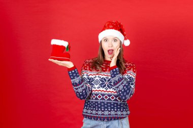  Elinde kar taneleri ve kırmızı arka planda Noel Baba şapkası olan bir kazakla elinde hediye ayakkabısı olan genç, şaşkın bir kadın.